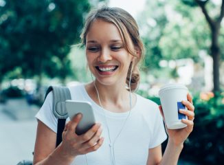 SEO Tips represented by closeup portrait of smiling young beautiful woman walking, holding drink, wearing headphones and having video call on smartphone on street